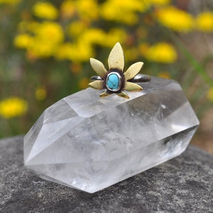 Pode incluir: Um anel de prata com um design de folha de ouro e uma pedra turquesa no centro. O anel está apoiado em um cristal de quartzo transparente.