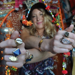 Puede incluir: Una mujer con un sombrero y un collar largo con cuentas y abalorios de colores. Lleva varios anillos en los dedos, incluyendo un anillo con una piedra azul y un anillo con una concha.