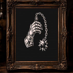 Könnte beinhalten: Ein gerahmter Druck mit einer weißen Illustration eines mittelalterlichen Panzerhandschuhs, der einen stacheligen Streitkolben auf schwarzem Hintergrund hält. Der verzierte, dunkelbraune Rahmen verleiht eine Vintage-Ästhetik. Der Druck wird auf einer Holzoberfläche präsentiert.