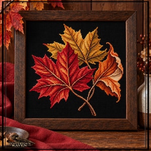 May include: A square cross-stitch embroidery featuring three overlapping autumn leaves in vibrant shades of red, orange, and gold, set against a solid black background. The artwork is encased in a dark brown wooden frame with a visible grain texture.