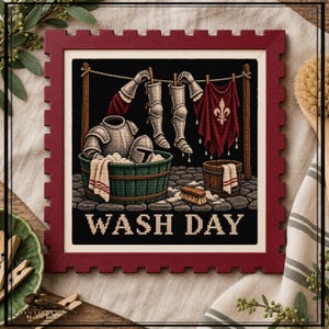 May include: A framed cross-stitch embroidery featuring a scene of medieval armour being washed in a green tub. The design includes a clothesline with metal greaves and a red banner with a fleur-de-lis, with the text WASH DAY at the bottom.