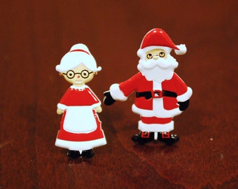 May include: Two Christmas figurines: Santa Claus and Mrs. Claus. Santa wears a red suit with black boots and a white beard. Mrs. Claus is in a red dress and white apron. Both have round glasses. Festive holiday decor.