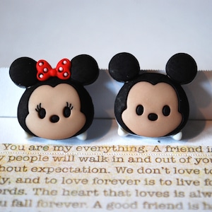 May include: Two small, cartoon-style figurines of Mickey Mouse and Minnie Mouse. Mickey is wearing black ears and a black and white face. Minnie is wearing black ears, a red bow, and a black and white face.