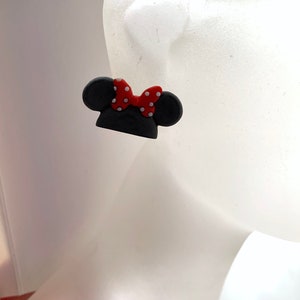 May include: A pair of black and red Minnie Mouse-inspired earrings. The earrings are shaped like Minnie Mouse's ears with a red bow with white polka dots.
