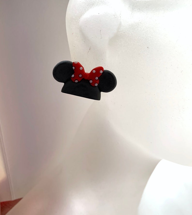 May include: A pair of black and red Minnie Mouse-inspired earrings. The earrings are shaped like Minnie Mouse's ears with a red bow with white polka dots.