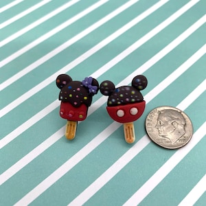 May include: Two Mickey Mouse-shaped popsicle charms. One has a purple bow, and both have chocolate-covered heads with colorful sprinkles. The popsicles are red with white accents and sit on wooden sticks. A dime is next to the charms.