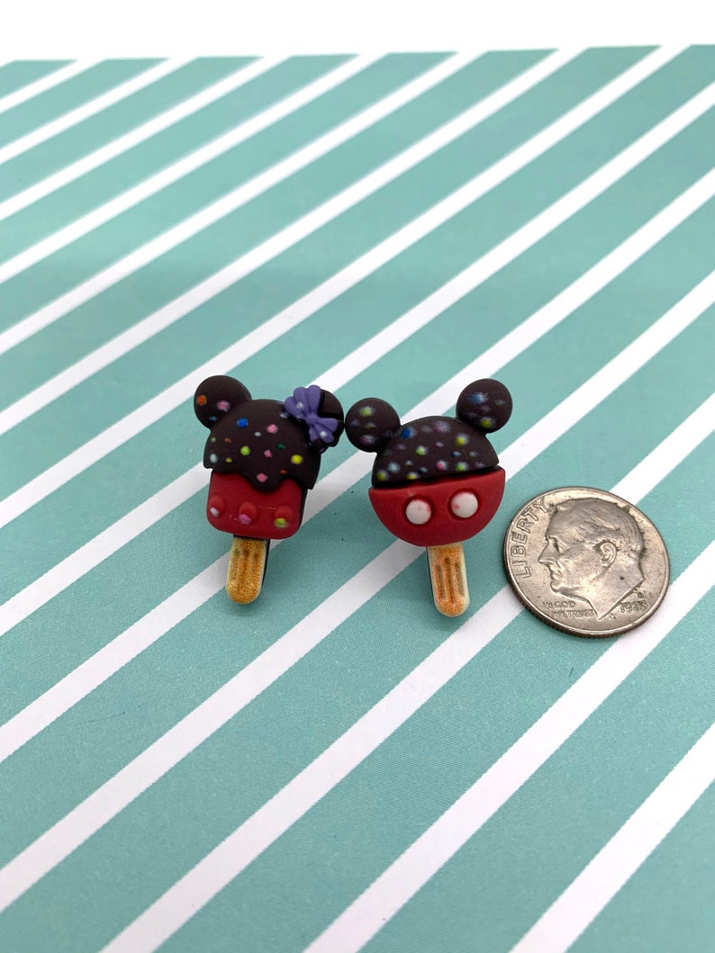 May include: Two Mickey Mouse-shaped popsicle charms. One has a purple bow, and both have chocolate-covered heads with colorful sprinkles. The popsicles are red with white accents and sit on wooden sticks. A dime is next to the charms.