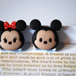 May include: A pair of small, cartoon-style figurines of Mickey Mouse and Minnie Mouse. Mickey is wearing black ears and a red nose. Minnie is wearing a red bow with white polka dots. Both figurines have brown eyes and a light brown face.