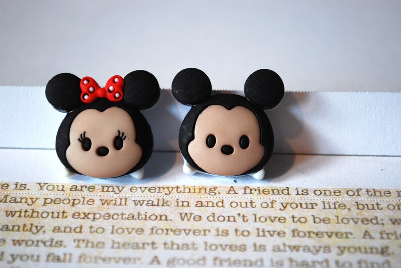May include: A pair of small, cartoon-style figurines of Mickey Mouse and Minnie Mouse. Mickey is wearing black ears and a red nose. Minnie is wearing a red bow with white polka dots. Both figurines have brown eyes and a light brown face.