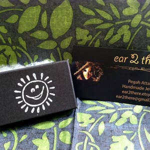 May include: A black box with a white smiling sun design. The box is sitting on a green and black patterned fabric. A business card with the text "ear 2 there" is visible in the background. The card includes the text "Pegah Attarpour Handmade Jewelry" and contact information.