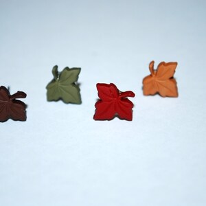May include: Four metal leaf-shaped pins in different colors: brown, green, red, and orange. The pins are arranged in a row on a white surface.