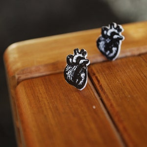 May include: Two black and white anatomical heart earrings on a wooden surface.