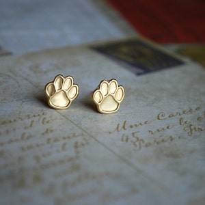 May include: Two gold-colored paw print stud earrings. The earrings are small, with a detailed paw print design. They are set against a vintage-style background with faded text.