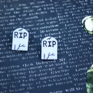 May include: Two white plastic tombstone-shaped charms with black lettering that reads "RIP". The charms are on a blue and white patterned surface.