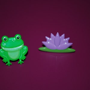 May include: A green frog figurine with a wide smile and a purple flower figurine with a green stem. Both figurines are made of plastic and have a glossy finish.