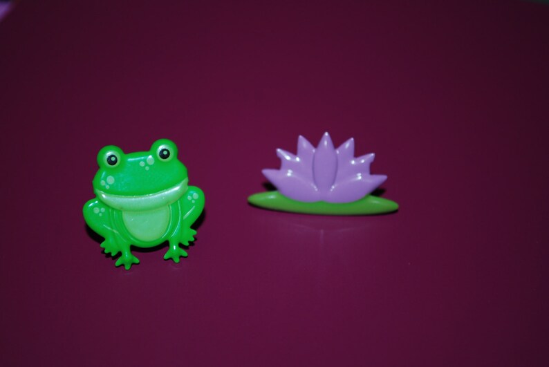 May include: A green frog figurine with a wide smile and a purple flower figurine with a green stem. Both figurines are made of plastic and have a glossy finish.