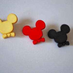 May include: Three small, cartoon-style Mickey Mouse heads with bow ties. The heads are yellow, red, and black.