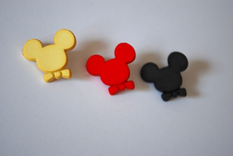 May include: Three small, cartoon-style Mickey Mouse heads with bow ties. The heads are yellow, red, and black.