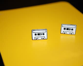 May include: Two white cassette tape pins on a yellow background. The pins are black and white and have a simple design.