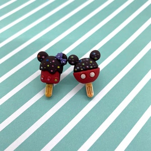 May include: Two Mickey Mouse-shaped popsicle charms. One is dark brown with colorful sprinkles and a purple bow, the other is red with black sprinkles. Both have wooden sticks and are on a striped background.