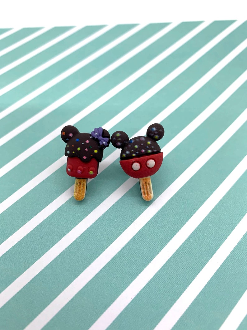 May include: Two Mickey Mouse-shaped popsicle charms. One is dark brown with colorful sprinkles and a purple bow, the other is red with black sprinkles. Both have wooden sticks and are on a striped background.