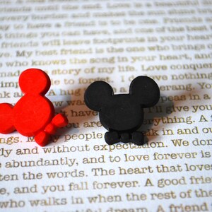 May include: Two small, cartoon-style Mickey Mouse figures. One is red and the other is black. Both figures have a bow tie and are sitting on a white surface with text.