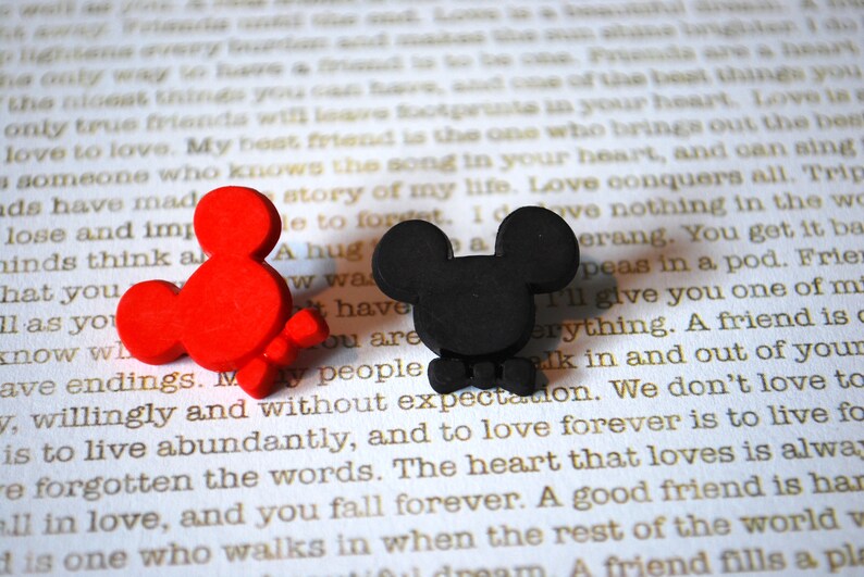 May include: Two small, cartoon-style Mickey Mouse figures. One is red and the other is black. Both figures have a bow tie and are sitting on a white surface with text.