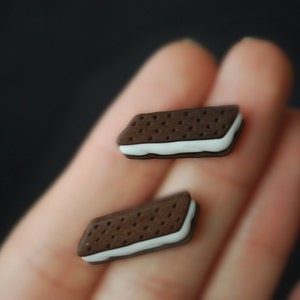 May include: Two miniature chocolate sandwich cookies with white filling. The cookies are held in a person's hand.