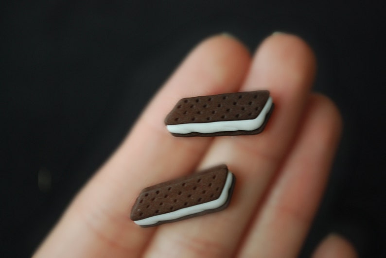 May include: Two miniature chocolate sandwich cookies with white filling. The cookies are held in a person's hand.
