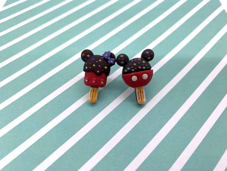 May include: Two Mickey Mouse-shaped ice cream popsicles. One is red with a purple bow, the other is red with a black head. Both have brown heads with colorful sprinkles and wooden sticks. The popsicles are on a light blue and white striped background.