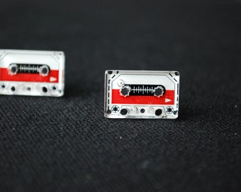 May include: Two retro cassette tape-shaped earrings. Each earring features a white and red design with black details, including the tape reels and a central sound level indicator. The earrings are set against a dark background.