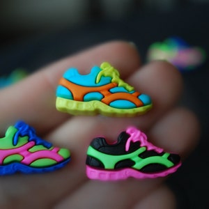 May include: Four colorful plastic shoe charms in different colors: green, blue, pink, and black. Each charm features a different color combination and a white shoelace detail.