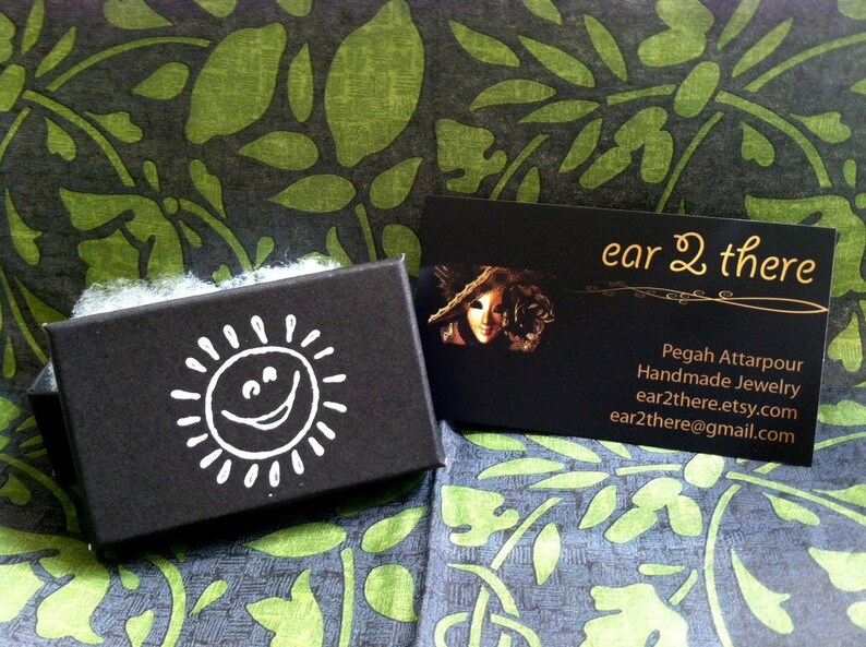 May include: A black box with a white smiling sun design. The box is sitting on a green and black patterned fabric. A business card with the text "ear 2 there" is visible in the background. The card includes the text "Pegah Attarpour Handmade Jewelry" and contact information.