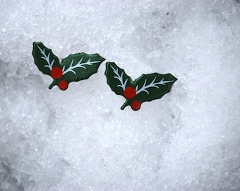 May include: Two green holly leaf ornaments with red berries on a white snowy background.