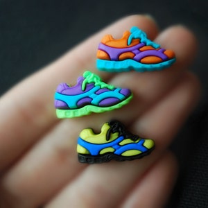 May include: Three colorful plastic shoe charms in the shape of running shoes. The shoes are green, blue, purple, orange, and yellow.