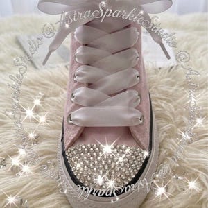 Puede incluir: Zapatilla alta rosa con cordones de satén blanco y un lazo grande. La puntera está adornada con brillantes pedrería. La zapatilla tiene una suela blanca con una franja negra. El texto en la imagen dice "Astra Sparkle Studio".