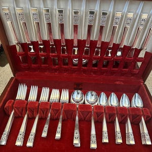 May include: A wooden silverware case with a red velvet interior holds a complete set of silver utensils. The set includes forks, knives, and spoons, each with ornate handle detailing. The case is lined with white satin.