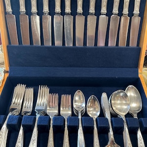 Könnte beinhalten: Ein silbernes Besteckset, bestehend aus Messern, Gabeln und Löffeln, angeordnet in einer Holzkiste mit blauem Samtinnenraum. Das Besteck hat dekorative Blumenmuster auf den Griffen.