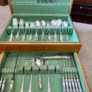 May include: A wooden cutlery chest with two drawers, lined with green felt. The drawers are filled with ornate silver-coloured forks, spoons, knives, and serving utensils. The cutlery has detailed floral patterns.
