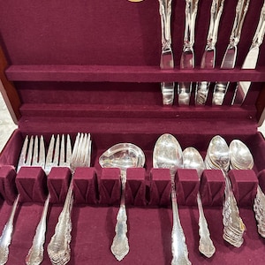 May include: A Naken tarnish proof silverware chest with a deep burgundy velvet interior. The set includes silver-toned metal forks, spoons, and knives arranged in dedicated slots. The handles feature an ornate, embossed floral pattern.