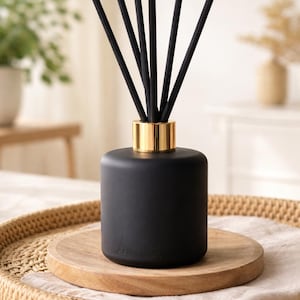 Op de afbeelding: Een zwarte etherische olie diffuser met een goudkleurige kraag en zwarte stokjes. De diffuser staat op een houten onderzetter en een gevlochten dienblad. Ontworpen voor huisgeur.