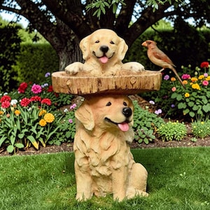 Puede incluir: Una estatua de jardín caprichosa con dos esculturas de golden retrievers. Un perro sostiene una plataforma con aspecto de madera con otro perro asomándose por encima. Un pequeño pájaro está posado cerca. La estatua está en un jardín exuberante.