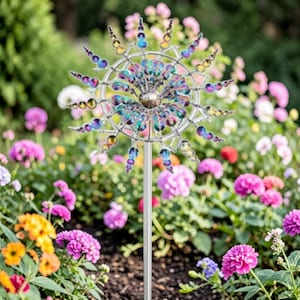 Puede incluir: Un molinillo de jardín decorativo de metal con detalles iridiscentes azules, morados y dorados. El molinillo está montado sobre un poste plateado y colocado en un jardín lleno de flores coloridas, incluyendo flores rosas, naranjas y moradas.