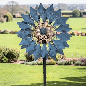 May include: A blue and gold metal garden spinner with a starburst design. The spinner has multiple blue star-shaped blades surrounding a central gold-colored floral design. It is mounted on a black metal pole and set in a grassy yard.