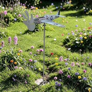 May include: A metal airplane wind spinner with a propeller and wings, mounted on a pole. The airplane is painted in light blue and white with black and white checkered accents. It is set in a garden with colorful flowers and green grass.