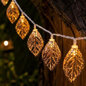 May include: A string of decorative lights featuring metallic gold-coloured leaf-shaped covers. Each leaf has a detailed, open-work vein pattern that allows the warm white light from the internal bulb to shine through, creating a glowing effect.