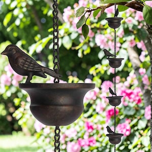 May include: A decorative bird feeder with a bronze finish. The feeder features a detailed bird perched on top of a bowl-shaped container, suspended by a chain. Additional smaller feeders with birds hang below. The background includes pink flowers and green foliage.