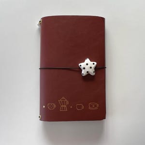 Könnte beinhalten: Ein weinrotes Lederjournal mit einem weißen sternförmigen Verschluss mit schwarzen Tupfen. Das Journal hat einen braunen elastischen Verschluss und goldfarbene Reißverschlüsse und Eckakzente. Goldfarbene Illustrationen zum Thema Kaffee sind auf der Vorderseite aufgedruckt.