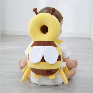 Puede incluir: Un protector de cabeza para bebé con forma de abeja amarilla y marrón. El protector de cabeza tiene un reposacabezas circular, rayas marrones, alas blancas y correas amarillas. Diseñado para proteger la cabeza del bebé mientras aprende a sentarse o gatear.
