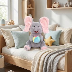 May include: A gray plush elephant toy with large pink ears and a smiling face. The elephant is holding a colorful ball and is sitting on a bed with pillows and a blanket. The toy is designed for children.